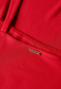 Czerwony materiałowy ubranie o gładkiej fakturze i subtelnym połysku. Zawiera małą mosiężną metkę "SUPERDRY". Przeszycia są widoczne na całej powierzchni.