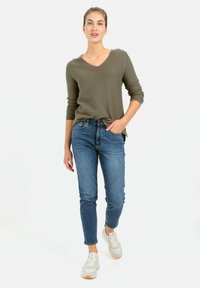 Oliwkowy sweter w serek z długimi rękawami, zestawiony z niebieskimi jeansami o slim fit oraz jasnoszarymi trampkami z białą podeszwą.