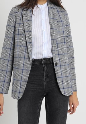 Mujer con blazer gris de cuadros sobre una camisa blanca abotonada y vaqueros negros de talle alto, de pie frente a un fondo liso.