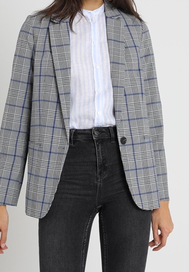 Femme portant un blazer à carreaux gris sur une chemise blanche boutonnée et un jean noir taille haute, debout devant un fond uni.