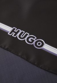 Zwarte stof met een opvallend "HUGO"-logo in het wit en paars, voorzien van een wit en paarse gestreepte rand.