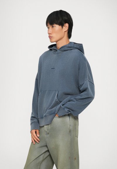 Felpa in cotone blu con tasca frontale, vestibilità comoda e polsini a coste. Indossata con pantaloni verde chiaro che presentano leggere variazioni di colore.