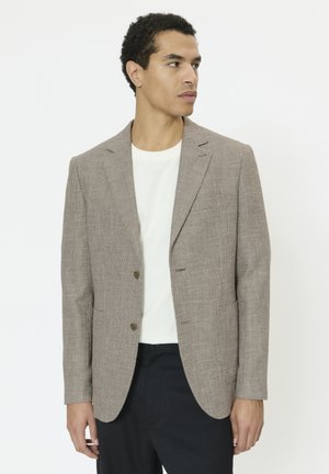 Mand iført en tekstureret lysebrun blazer over hvid skjorte og sorte bukser, der kigger til siden mod en ensartet hvid baggrund.