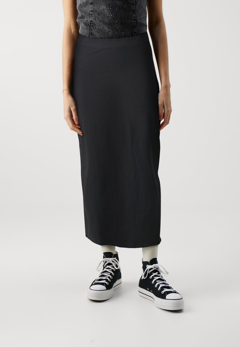 Femme portant un haut noir sans manches, une longue jupe noire, des chaussettes blanches et des baskets montantes noires, debout devant un fond uni.
