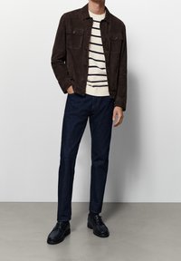 Braune Wildlederjacke über einem cremegestreiften Strickpullover, kombiniert mit dunkelblauen Jeans und schwarzen Schnürschuhen. Sanfte Texturen, lässige Passform.