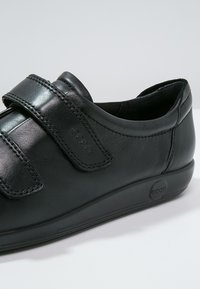Scarpa nera in pelle con una superficie liscia, dotata di una ampia fascia in velcro e una suola bassa e imbottita. Logo impresso sul lato.