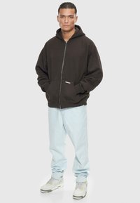Schwarze Zip-Hoodie mit oversized Schnitt, Fronttaschen und strukturiertem Stoff. Kombiniert mit hellblauen Jeans und weißen Sneakern.