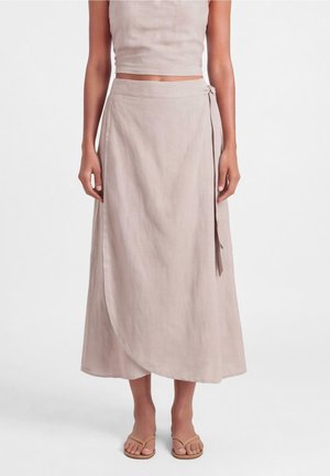 Vrouw draagt een lichtbeige wikkelrok gestrikt in de taille, gecombineerd met een bijpassende cropped top en platte sandalen tegen een witte achtergrond.
