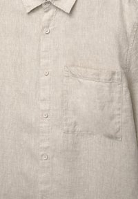 Gros plan sur le devant d'une chemise en lin beige avec des boutons au centre et une poche carrée sur la poitrine, côté gauche.