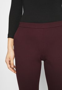 Leggings stretch bordeaux avec une texture lisse et une taille élastique, présentant un design minimaliste et sans coutures visibles.
