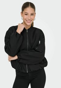 Blouson aviateur noir avec une fermeture éclair à l'avant, des poignets élastiqués et une poche zippée sur la manche. Le tissu a une finition lisse.
