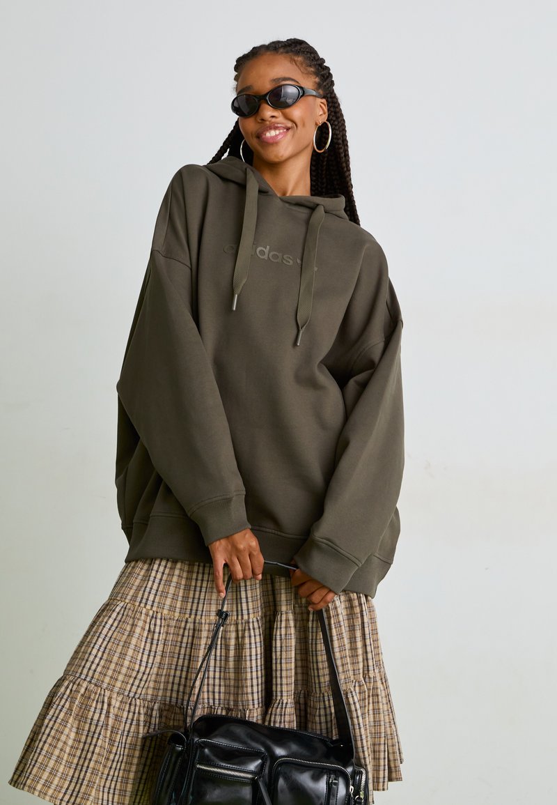 Olivgrön oversized hoodie med dragsko, med en subtil logotyp. Kombinerad med en skiktad rutkjol och en svart väska.