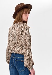 Kobieta w brązowym kapeluszu, bluzce z długim rękawem w panterkę oraz niebieskich jeansach, patrząca na bok na jednolitym białym tle.