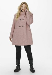 Manteau double boutonnage rose clair avec une finition texturée, boutons noirs, grandes poches et un col haut, porté sur un pantalon noir et des bottes.