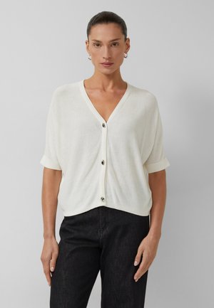 Frau trägt eine weiße kurzärmelige, geknöpfte Strickjacke mit V-Ausschnitt und schwarze Hose, steht vor einem schlichten grauen Hintergrund.