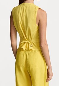 Žlutý top bez rukávů s přiléhavým střihem, detaily na zavázání na zádech a texturovanou látkou. V kombinaci se žlutými kalhotami se zadními kapsami.