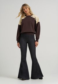 Καφέ φούτερ με κρεντίζ (κρέμα) σχέδιο φύλλου και ριμπ μανσέτες, συνδυασμένο με τζιν παντελόνι σε στυλ καμπάνα από σκούρο τζιν, με κάθετη υφή και διακριτική γυαλάδα.