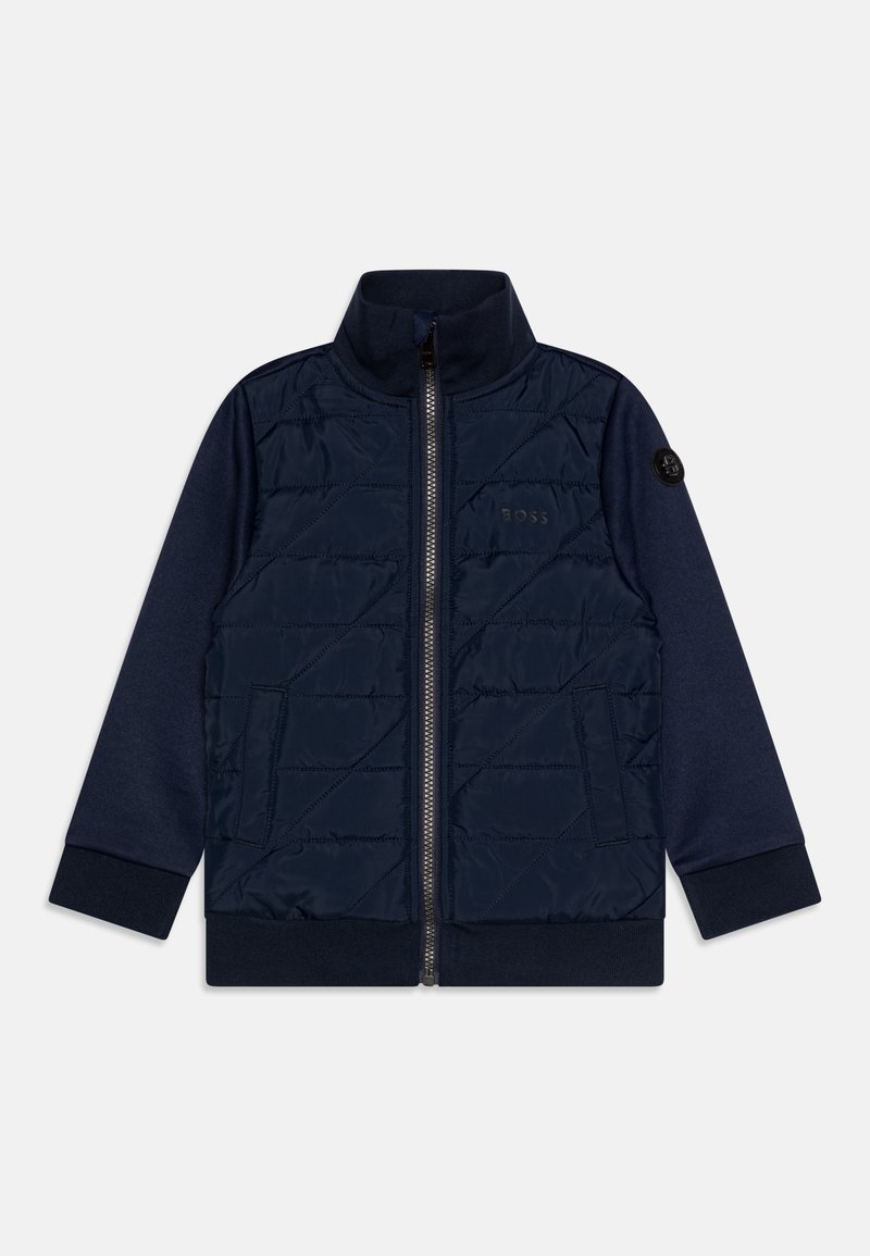 Veste bleu marine avec un devant matelassé, col haut, fermeture éclair sur toute la longueur et poignets côtelés ; présente un écusson logo sur la manche et des poches latérales.