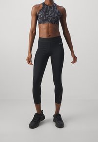Vältränad person i svart mönstrad sportbh, svarta cropped leggings med vit Nike-logotyp och svarta sportskor som står på en grå golv.