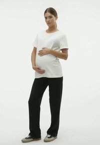 Pessoa gravida vestindo uma blusa branca de manga curta e calças pretas, de pé com uma mão na barriga e um fundo neutro.