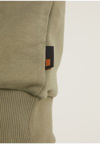 Felpa khaki con polsini a costine, dotata di un piccolo etichetta nera con accenti arancioni. Tessuto morbido con una texture liscia.