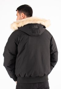 Veste noire avec une grande capuche bordée de fourrure. Présente des poignets et une ceinture côtelés, ainsi qu'un tissu extérieur lisse. Pas de marquage ni de motifs visibles.