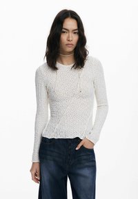 Top en dentelle blanche à manches longues avec un motif texturé, décolleté rond et détail à cordon, associé à un denim taille haute bleu foncé.