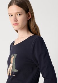Marineblaue Langarmbluse aus einem Baumwollmischgewebe mit einem Grafikdruck eines Hundes neben grünen Gummistiefeln. Rundhalsausschnitt, glatte Textur.