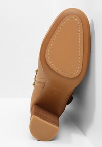 Semelle de chaussure en cuir brun clair avec un patch antidérapant cousu et un talon empilé en bois, vue par le dessous sur fond blanc.