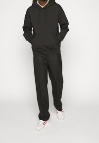 Černá mikina s kapucí a přední kapsou, spárovaná s volnými tmavými kalhotami a bílými teniskami s červenými akcenty.