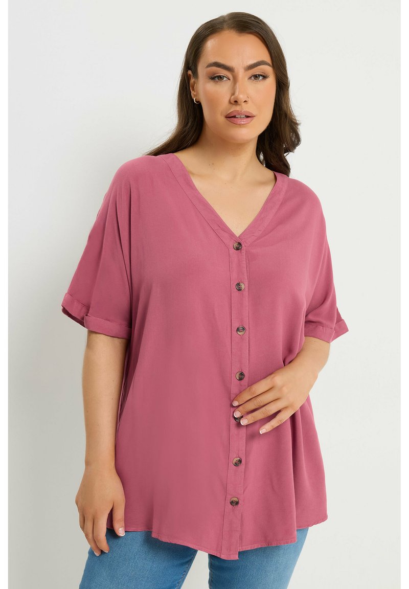 Donna che indossa una camicetta lilla, corta, a maniche corte, con bottoni e scollo a V, e jeans azzurri, in piedi davanti a uno sfondo chiaro e uniforme.