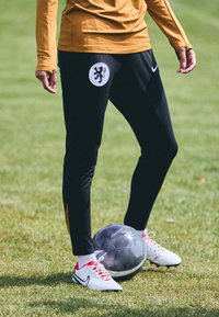 Svarte treningbukser med et lionlogo på en hvit lapp, paret med hvite og røde fotballsko, stående over en svart fotball på gress.
