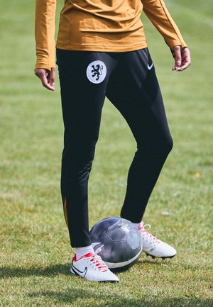 Černé sportovní kalhoty s logem lva na bílém výpletu, v kombinaci s bílými a červenými fotbalovými botami, postavené nad černým fotbalovým míčem na trávě.
