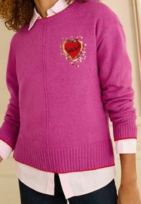 Rózsaszín kötött pulóver, szív alakú flitteres folttal, rajta a "Wish Love" felirattal. Bordázott szegéllyel és mandzsettákkal, fehér galléros ing fölé rétegezve.