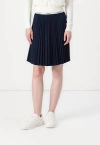 Navy pleated skirt with a white branded waistband, made of fabric, paired with white sneakers. The design features sharp pleats for texture.