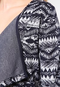 Námořnický cardigan s geometrickým bílým vzorem, texturovanou pleteninou a kontrastní šedou přední částí s čistým zakončením okrajů.