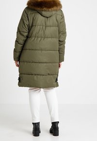 Langer olivgrüner Puffer-Mantel mit einer Kapuze mit Fellbesatz. Verfügt über ein gestepptes Design und verstellbare Kordeln am Saum. Trägt man über weißen Hose und schwarzen Stiefeln.