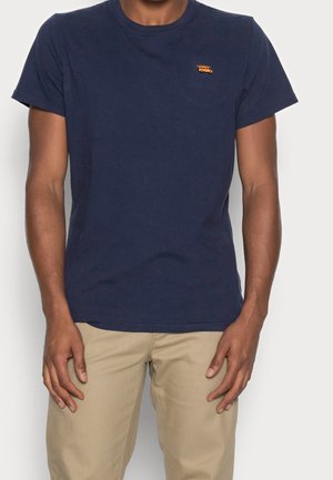 Homme portant un t-shirt à manches courtes bleu marine avec un petit logo de bus orange sur la poitrine gauche, associé à un pantalon beige.