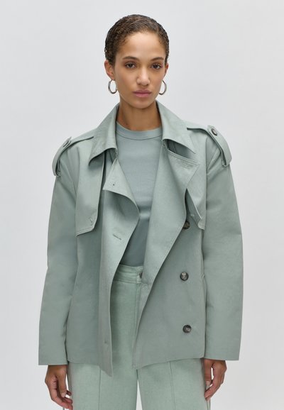 Γυναίκα με κοντά σγουρά μαλλιά φοράει πράσινο ανοιχτόχρωμο παλτό μέσης, ταιριαστό παντελόνι ψηλής μέσης και ριμπ τοπ με λαιμόκοψη, φόντο γκρι μονόχρωμο.