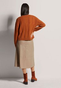 Gebreide roestkleurige trui, beige leren midirok en bruine enkellaarzen met een blokhak. De gestructureerde trui heeft een kabelgebreid ontwerp.