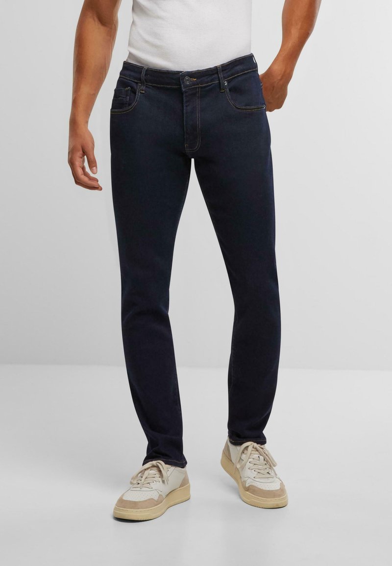 Tumši zilo džinsu bikses ar šauru piegriezumu, kas aprīkotas ar pogu jostasvietā, piecām kabatām un kontrastējošām dzeltenām šuvēm. Parādītas uz neitrāla fona.