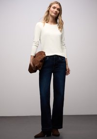 Hvid langærmet top med rund hals, kombineret med mørkeblå jeans. En brun ruskindstaske holdt i den ene hånd. Mørkebrune ankelstøvler.