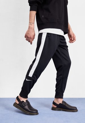 Person iført sorte Nike træningsbukser med hvide sidelister og sorte lædersandaler med spændere, stående på en blå gulv.
