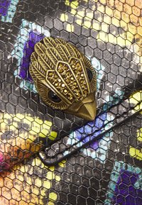 Barevný materiál se vzorem hada s hexagonální strukturou, který má zlatou sponu ve tvaru ptáka zdobenou rhinestones a černýma očima.