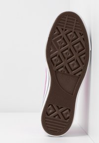 Brun gummisula med ett diamantmönster. Har en vit kant och präglad "Converse All Star"-logotyp. Rosa sidopanel synlig.