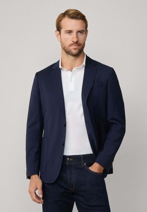 Marineblazer mit zwei Knöpfen, Reverskragen und einer Brusttasche, getragen über einem weißen Poloshirt und dunklen Jeans. Glattes Material und taillierte Passform.
