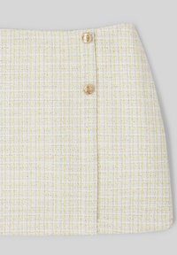 Jupe en tweed blanche et jaune avec une finition texturée, comportant deux boutons dorés à l'avant pour l'accent. Forme rectangulaire, longueur au-dessus du genou.