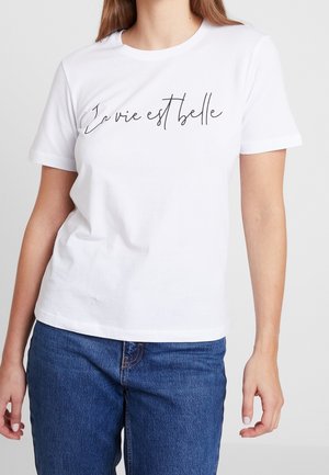 Kvinde iført hvid T-shirt med korte ærmer med teksten "La vie est belle" i sort skrift, kombineret med blå cowboybukser, stående foran en ensfarvet baggrund.