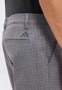 Κοντινό πλάνο ενός ατόμου που φορά γκρι καρό παντελόνια με ένα μαύρο λογότυπο κοντά στην πίσω τσέπη και ένα μαύρο πουκάμισο με σχέδια μέσα στην ζώνη.