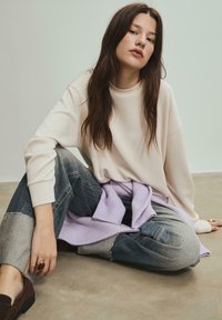 Creme Sweatshirt, lockere Passform, runder Ausschnitt. Kombiniert mit lässigen Patchwork-Jeans in Blau und Grau. Lila Strickpullover um die Taille gebunden.